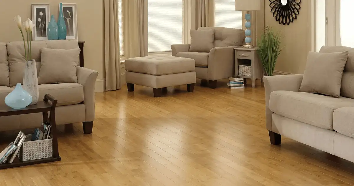 A living room with professionally installed bamboo flooring, beige sofas, a matching ottoman, a side table with a blue lamp, vases, framed pictures, and large windows with blinds and curtains in Green Bay, WI